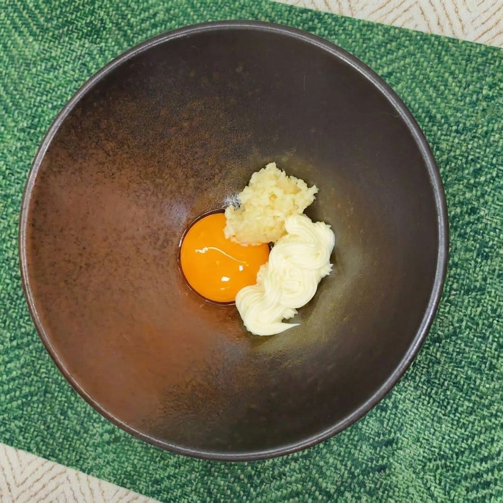 Un cuenco cerámico con una yema de huevo, mayonesa y ajo picado sobre un mantel individual verde.
