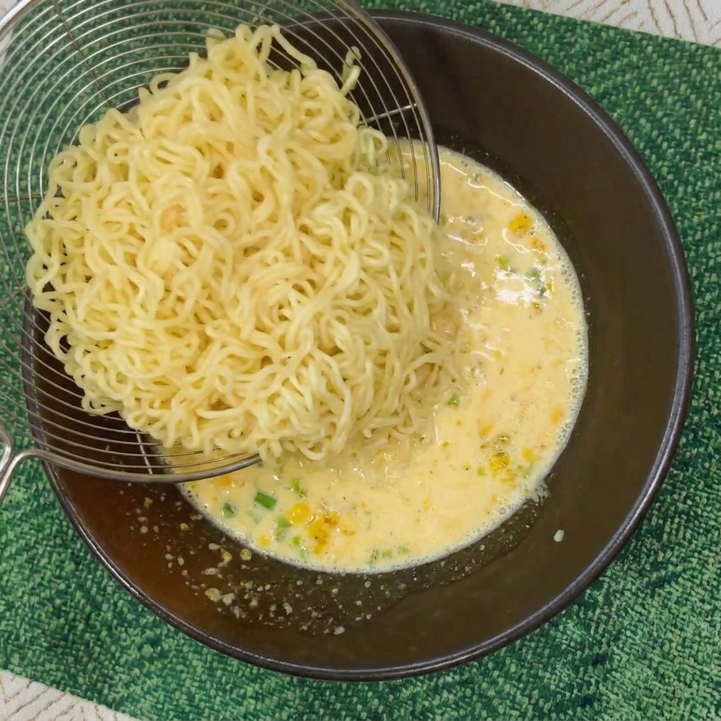 Un colador de metal transfiriendo fideos instantáneos rizados a un cuenco con caldo cremoso.