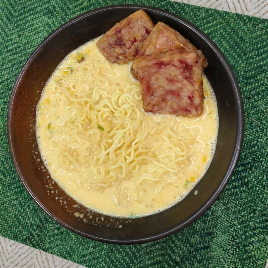 Fideos rizados en caldo cremoso cubiertos con tres lonchas de Spam doradas en un cuenco oscuro.