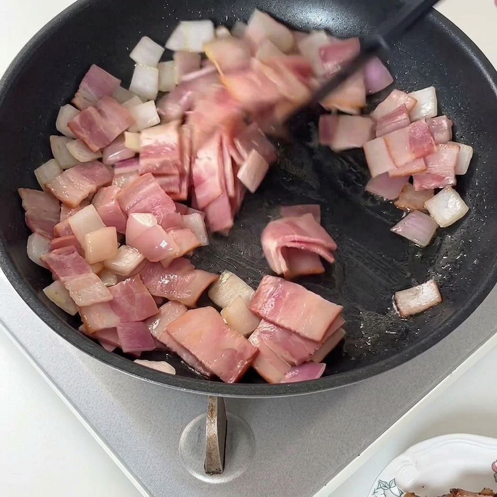 Una espátula removiendo el beicon cocinado y la cebolla en una sartén.