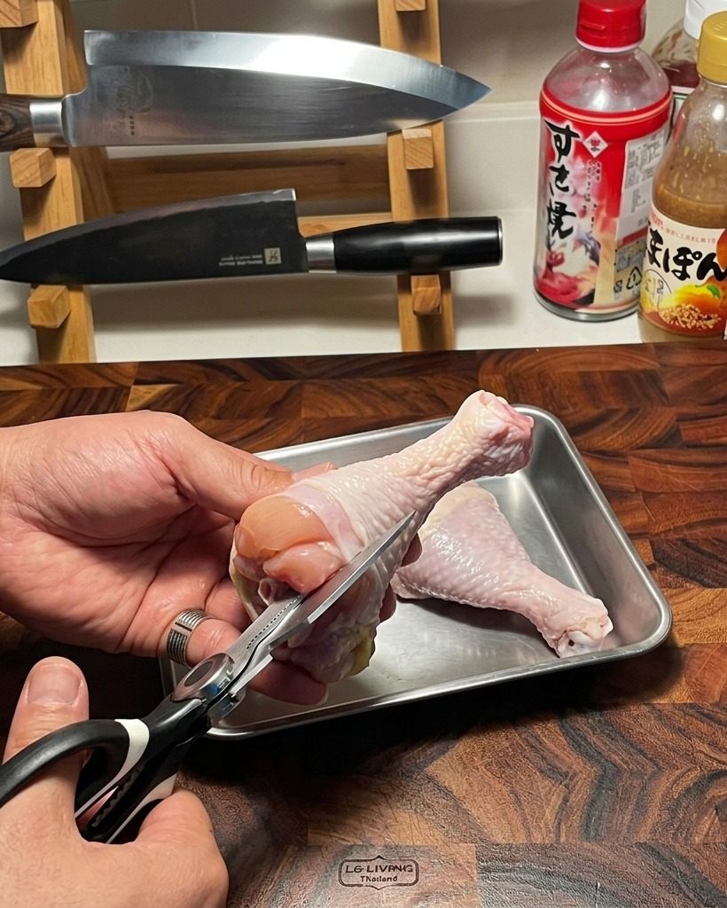 Unas tijeras de cocina retirando con cuidado el hueso de un contramuslo de pollo crudo sobre una tabla de cortar de madera.