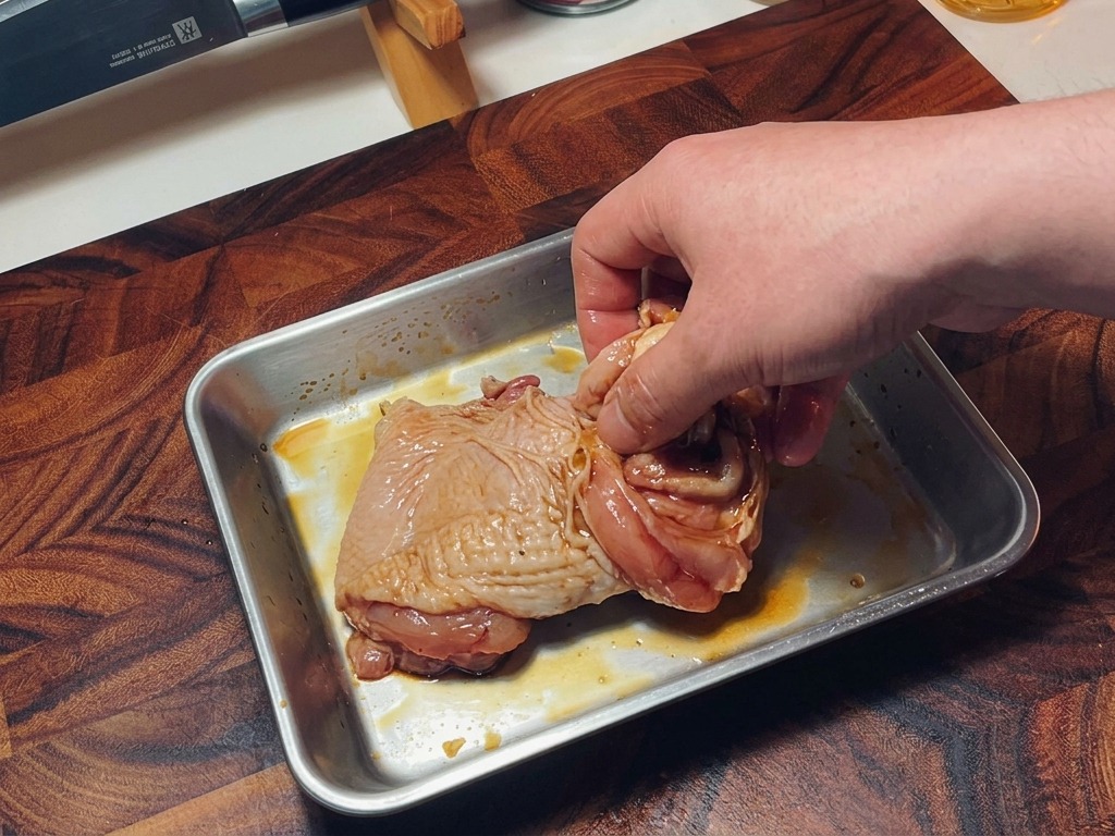 Una mano mezclando los contramuslos de pollo sazonados en una bandeja para asegurar que el marinado se distribuya bien.