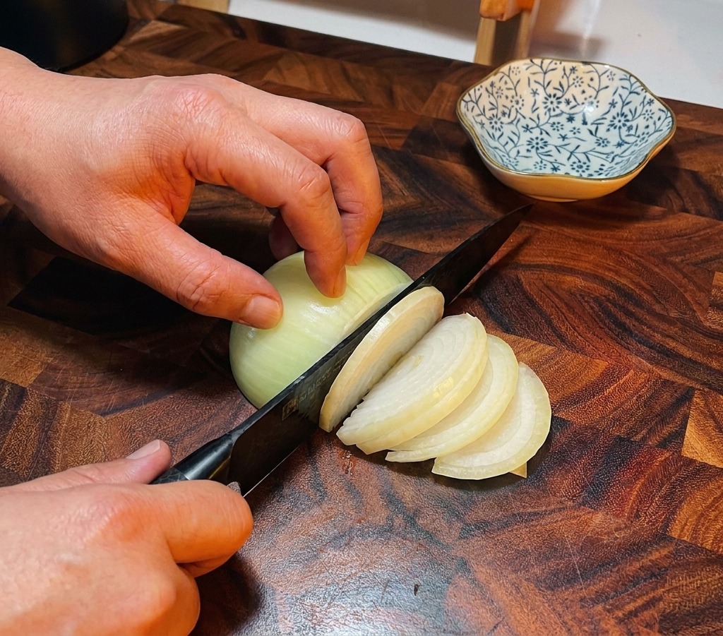Un cuchillo de chef rebanando una cebolla blanca en una tabla de cortar de madera oscura.