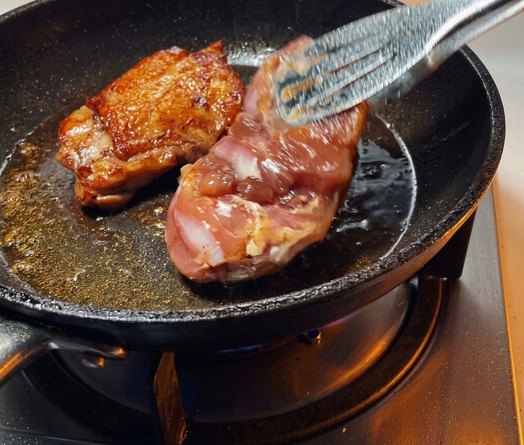Unas pinzas dando la vuelta a un contramuslo de pollo en la sartén, mostrando una piel perfectamente dorada y crujiente.