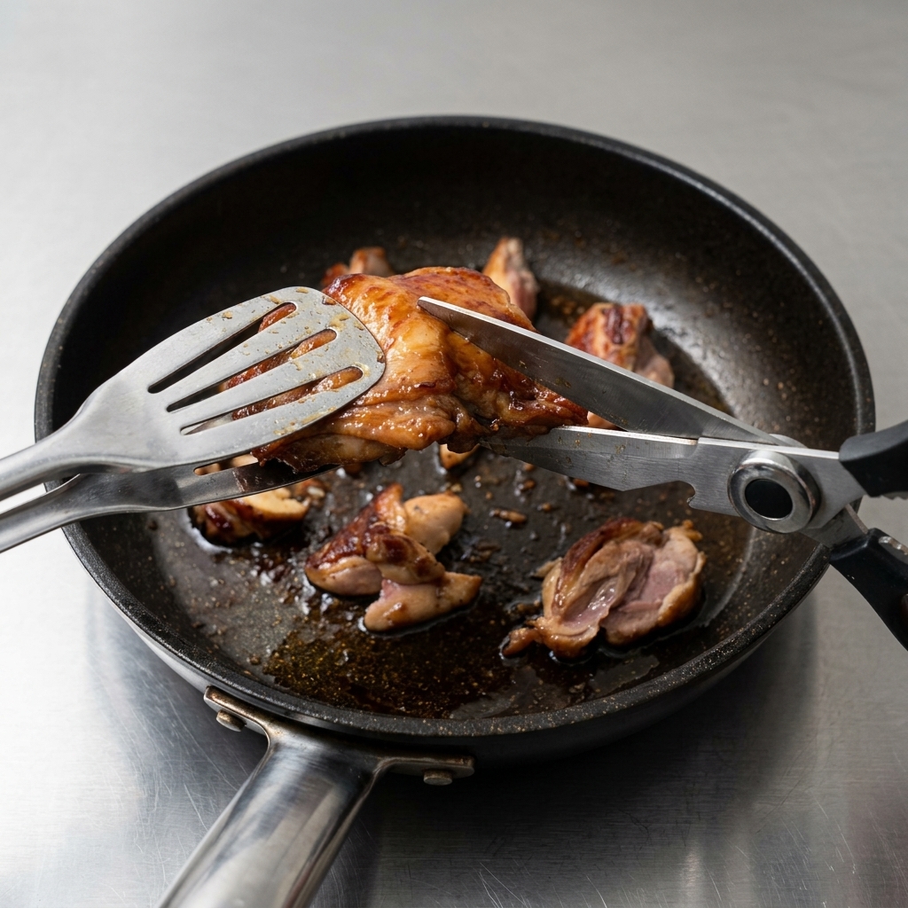 Unas tijeras de cocina cortando los contramuslos de pollo dorados directamente en la sartén en trozos pequeños.