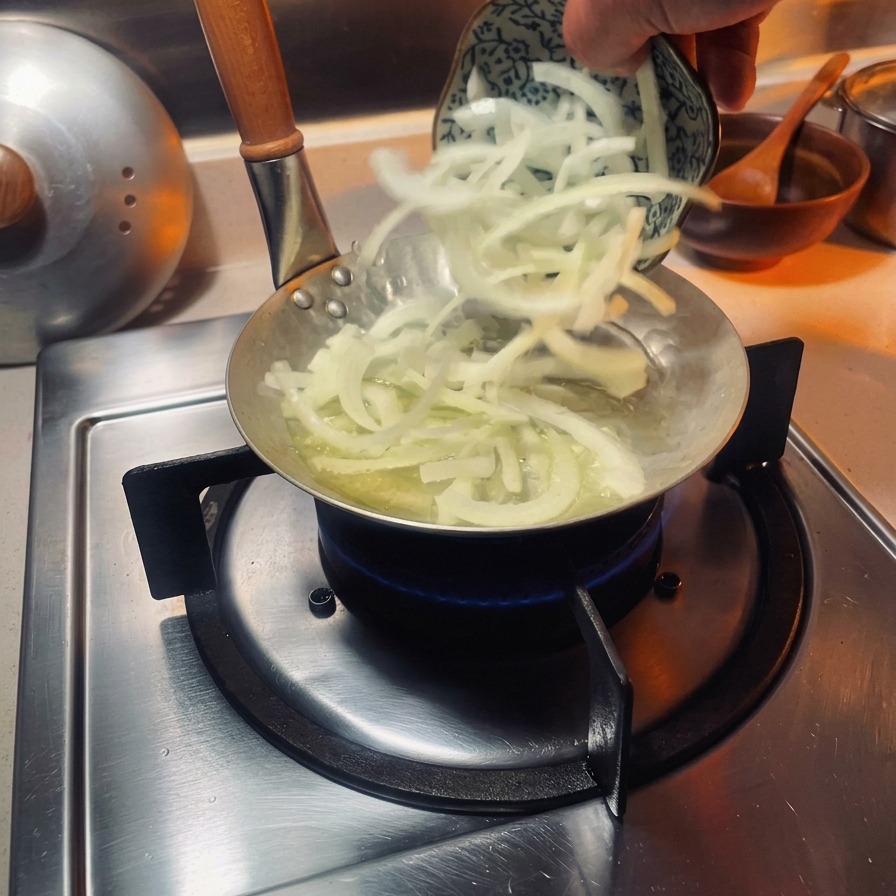 Cebolla cortada en juliana incorporándose a una sartén pequeña especial para oyakodon sobre un quemador de gas.