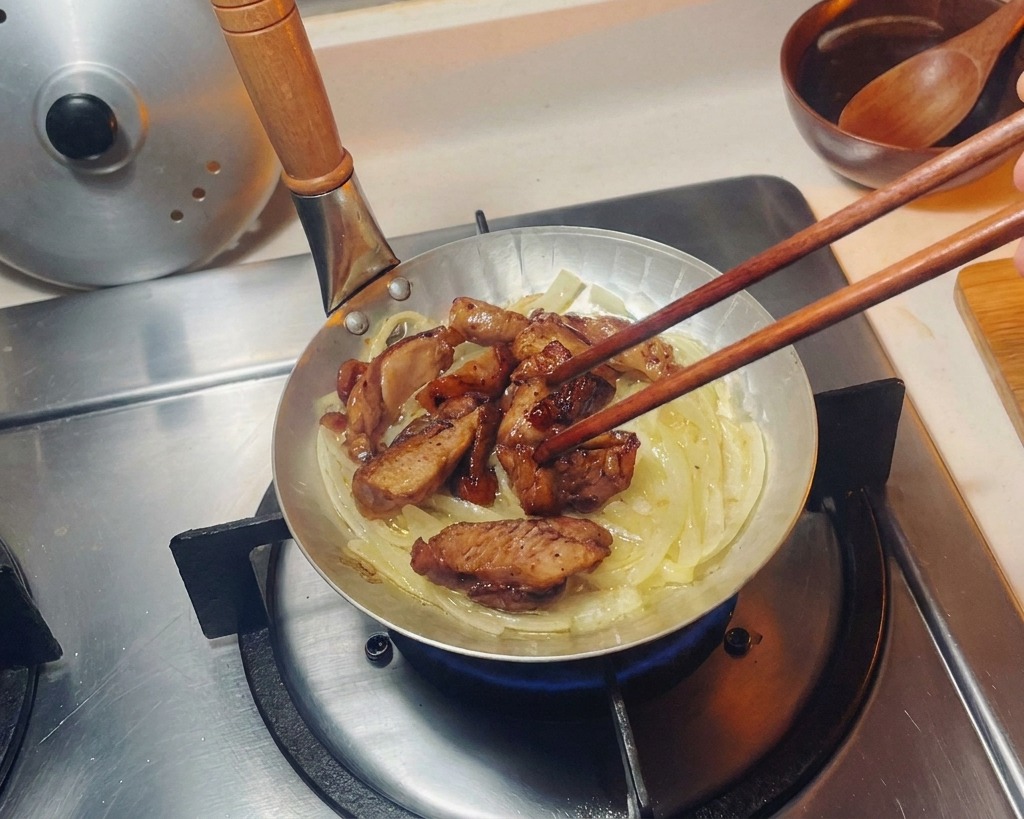Trozos de pollo dorado acomodándose con palillos sobre una cama de cebolla translúcida en una sartén pequeña.