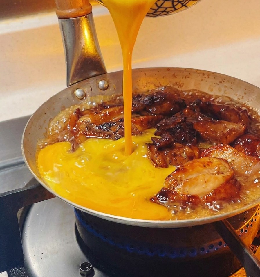 Huevo batido con tonos dorados vertiéndose en hilos circulares sobre el pollo que burbujea en la sartén.