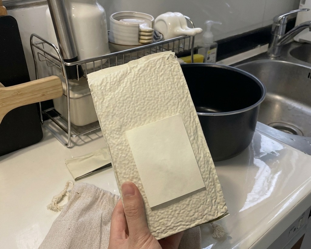 Una mano sosteniendo una bolsa blanca texturizada, vertiendo arroz blanco seco de grano corto en la cubeta interior negra de una arrocera sobre una encimera de cocina limpia.