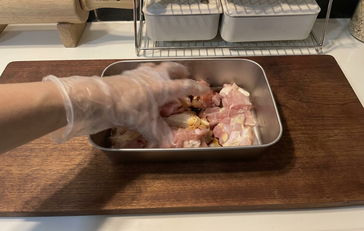 Una mano con un guante de plástico transparente mezclando trozos de pollo crudo con un marinado de salsa de soja oscura en una bandeja metálica poco profunda sobre una tabla de cortar de madera.