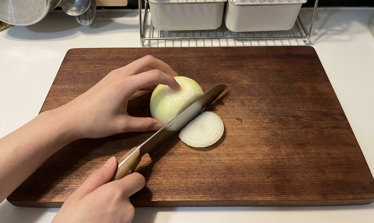 Manos utilizando un cuchillo con mango de madera para cortar una cebolla blanca en tiras finas de media luna sobre una tabla de cortar de madera oscura.