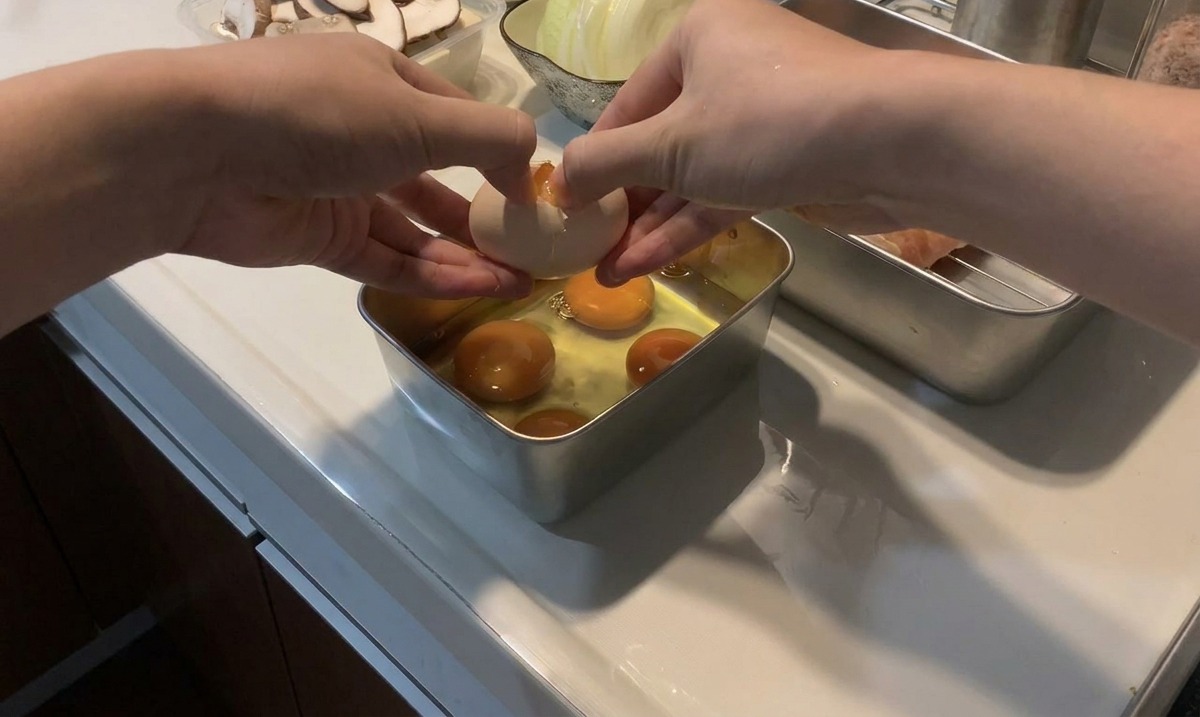 Manos cascando un huevo fresco sobre una bandeja metálica que ya contiene varias yemas y claras de huevo crudas.