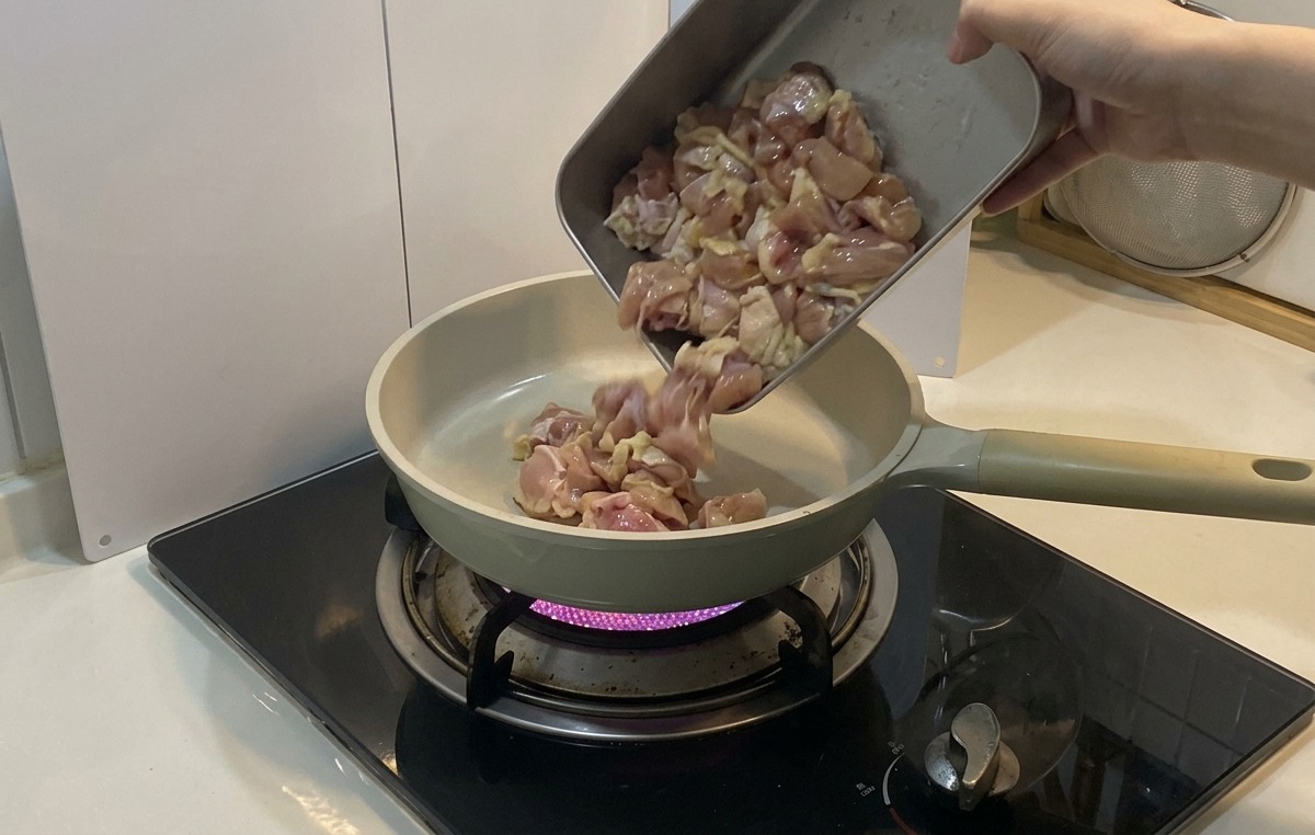 Trozos de pollo marinado vertiéndose desde una bandeja metálica en una sartén de cerámica de color claro sobre una llama de gas púrpura.