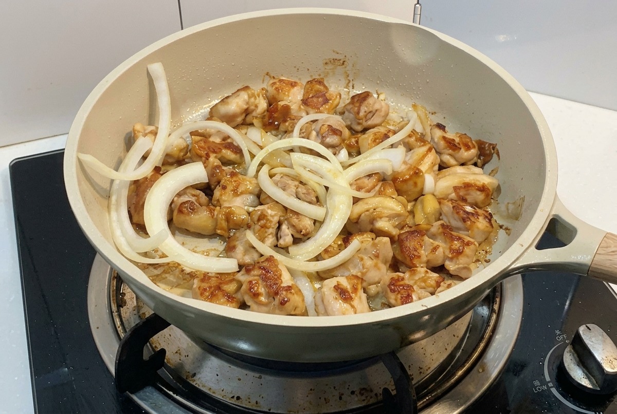 Aros de cebolla blanca laminada añadiéndose a una sartén con trozos de pollo que se están dorando en la cocina.