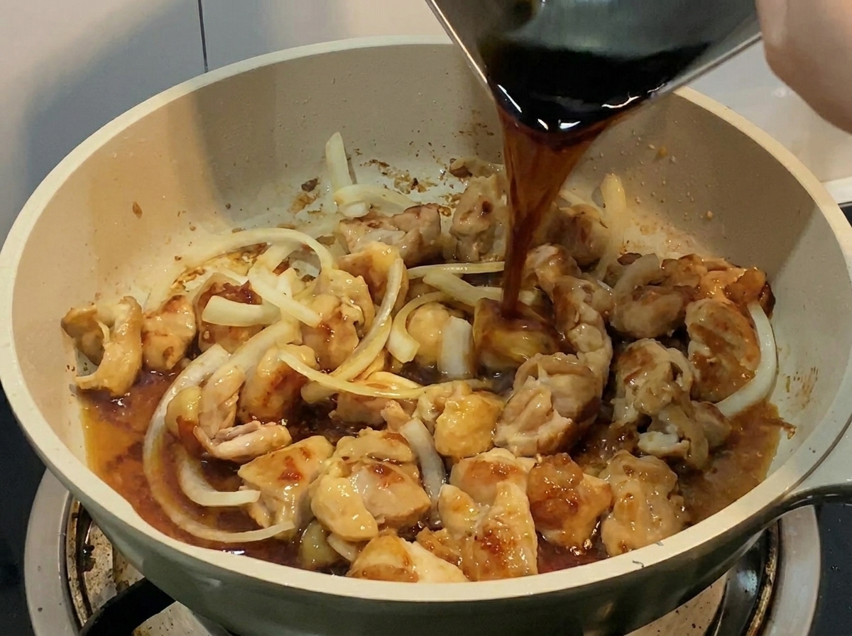 Un líquido oscuro y sabroso vertiéndose desde un recipiente metálico en una sartén llena de pollo y cebolla.