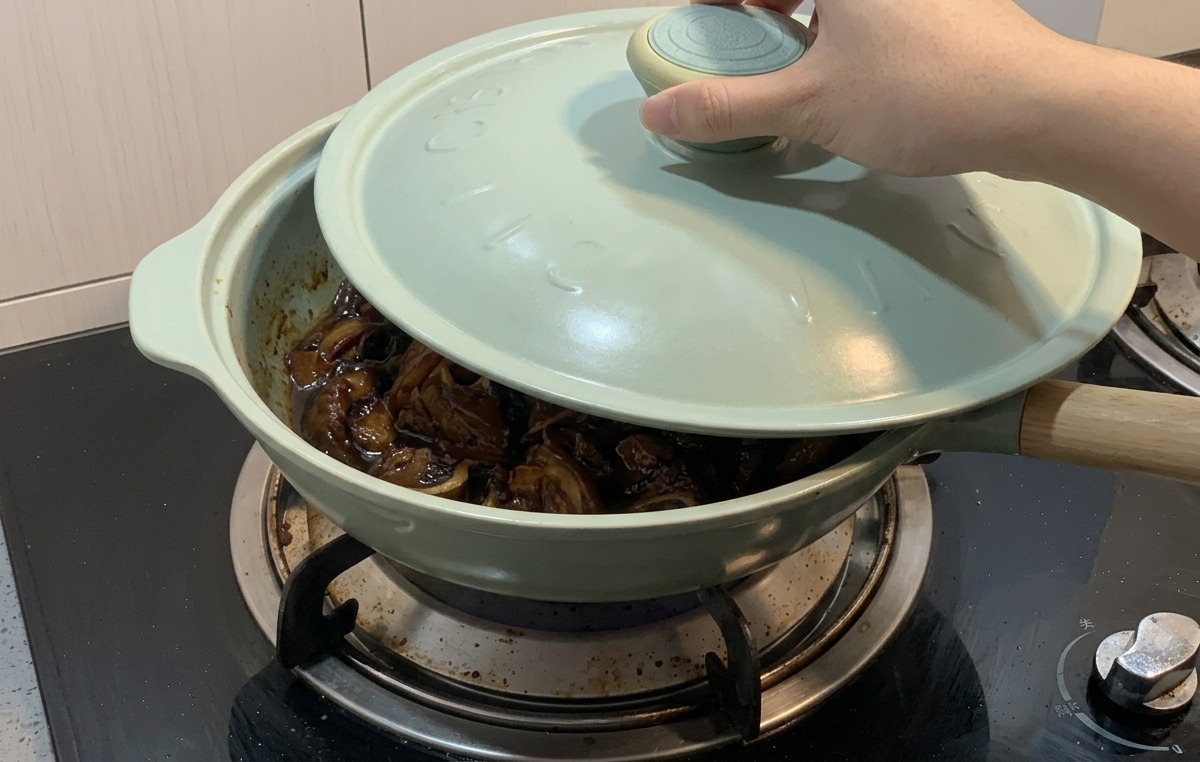 Una mano colocando una tapa azul claro sobre una sartén verde claro que contiene pollo y cebollas hirviendo a fuego lento en una salsa oscura.