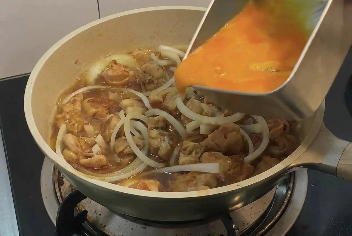 Una mano vertiendo huevo batido desde una bandeja metálica rectangular en una sartén con trozos de pollo y tiras de cebolla.