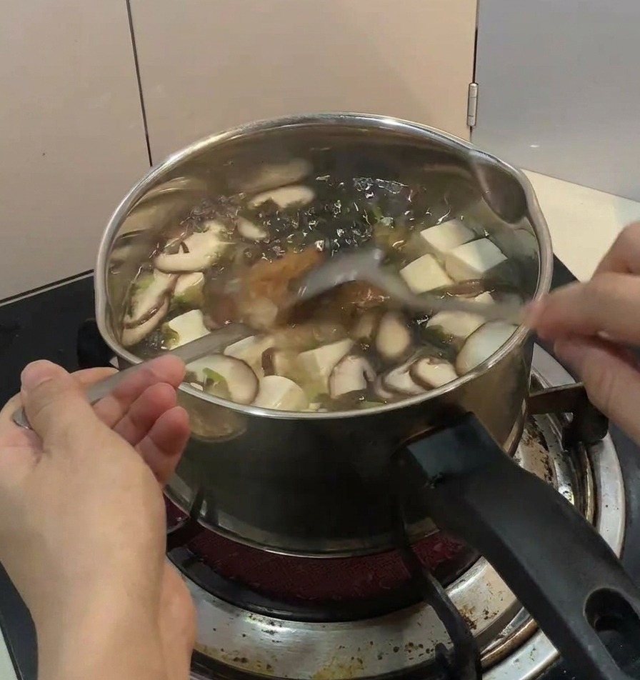 Una mano espolvoreando rodajas de cebollino fresco y tiras de alga nori oscura sobre un bowl humeante de arroz con pollo y huevo en una mesa de madera.
