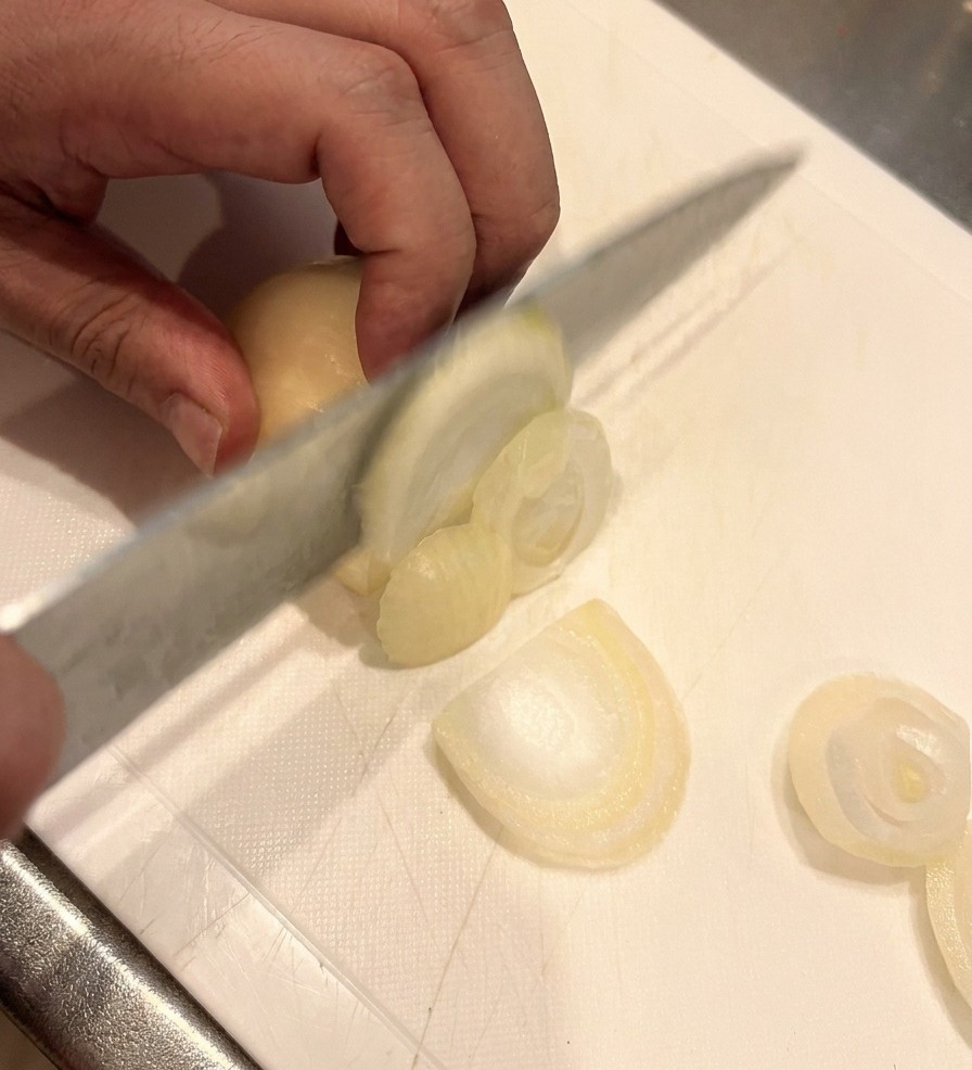 Un cuchillo afilado cortando una cebolla blanca en gajos finos sobre una tabla de cortar blanca, con una mano sosteniendo la cebolla de forma segura.