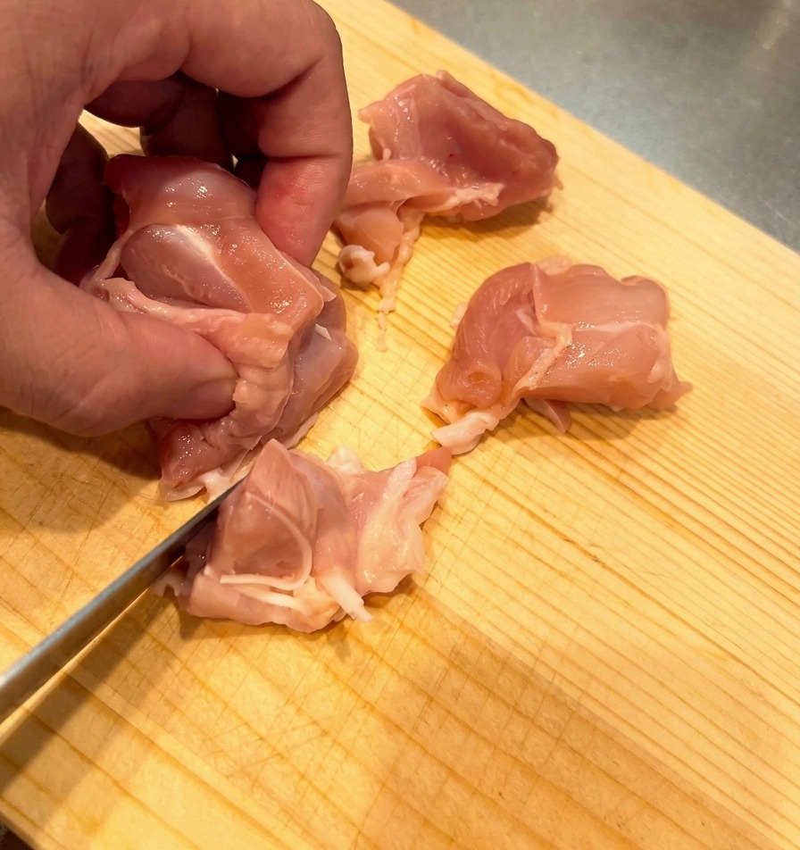 Carne cruda de contramuslo de pollo cortada en trozos pequeños y uniformes sobre una tabla de madera de color claro.