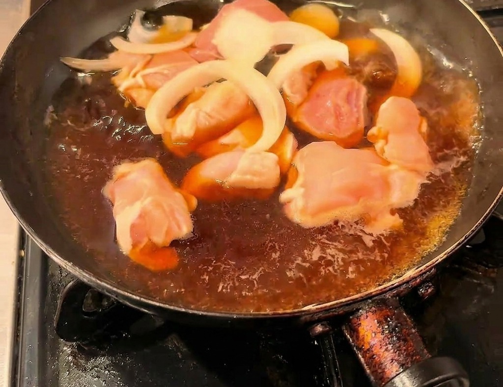 Trozos de pollo y cebolla en rodajas cocinándose a fuego lento en un caldo oscuro de salsa de soja dentro de una sartén negra.