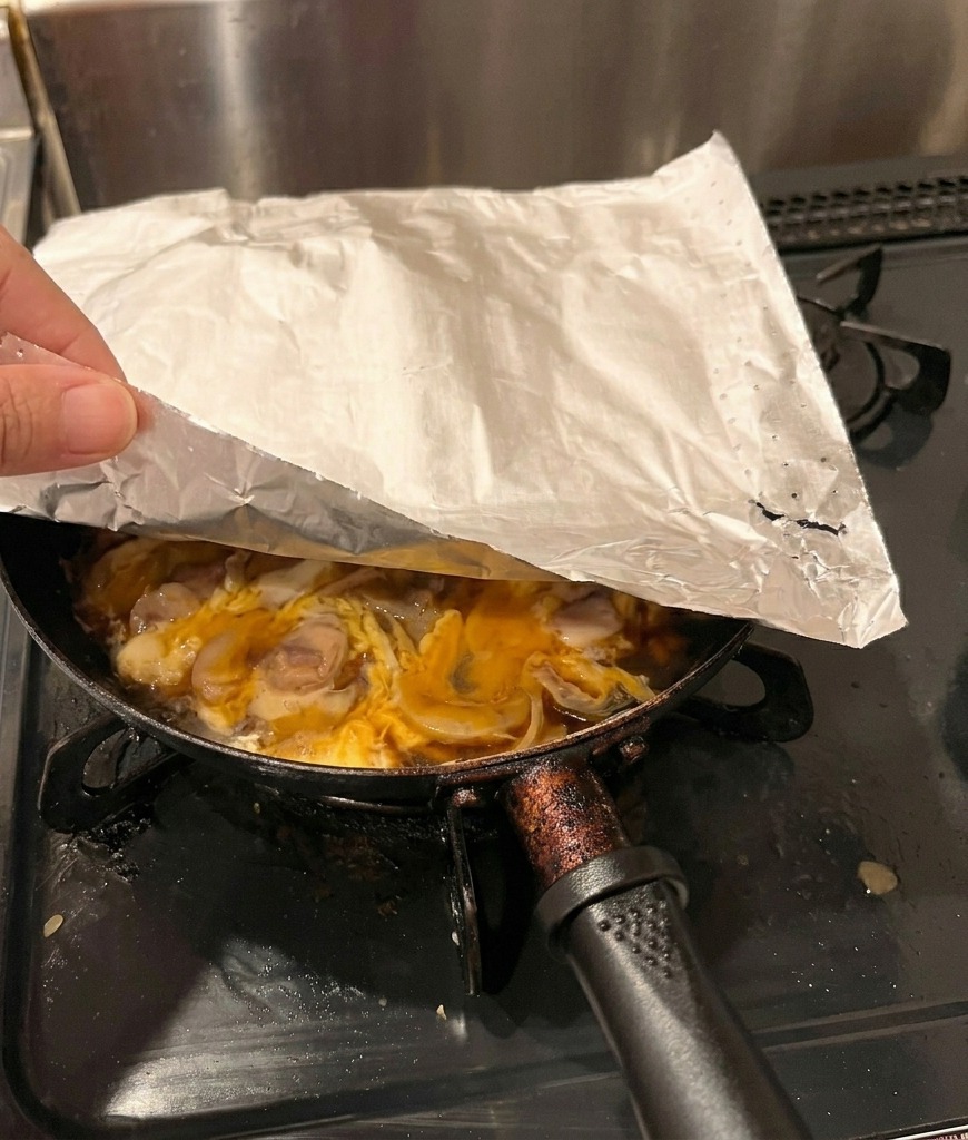 Una mano colocando una hoja de papel de horno blanco sobre una sartén con pollo y huevos parcialmente cocidos.