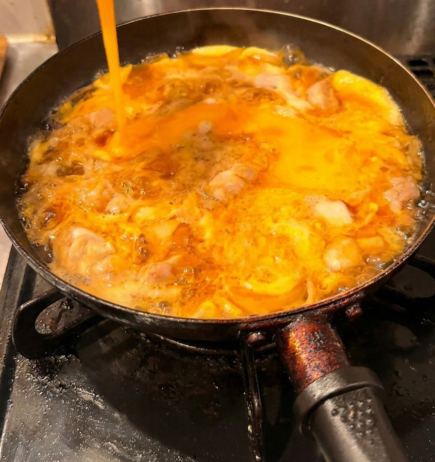 Un hilo de huevos batidos de color amarillo brillante vertiéndose en una sartén sobre el pollo y una capa base de huevos cocidos.