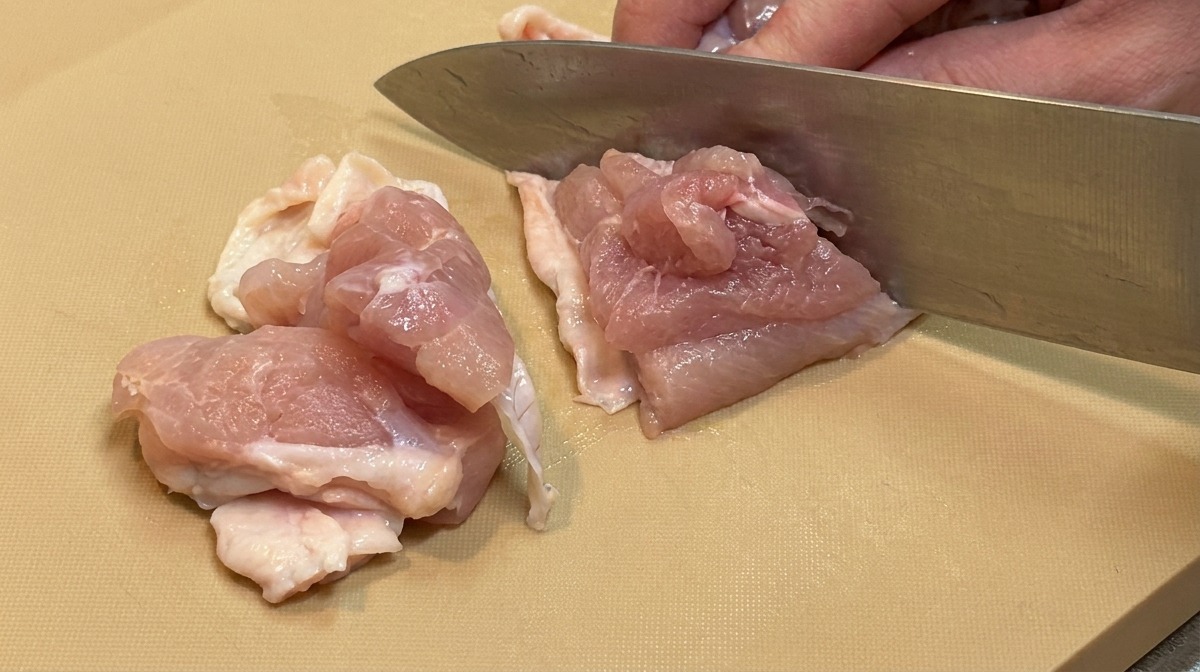 Carne cruda de contramuslo de pollo siendo cortada en trozos pequeños y uniformes con un cuchillo de cocina grande.
