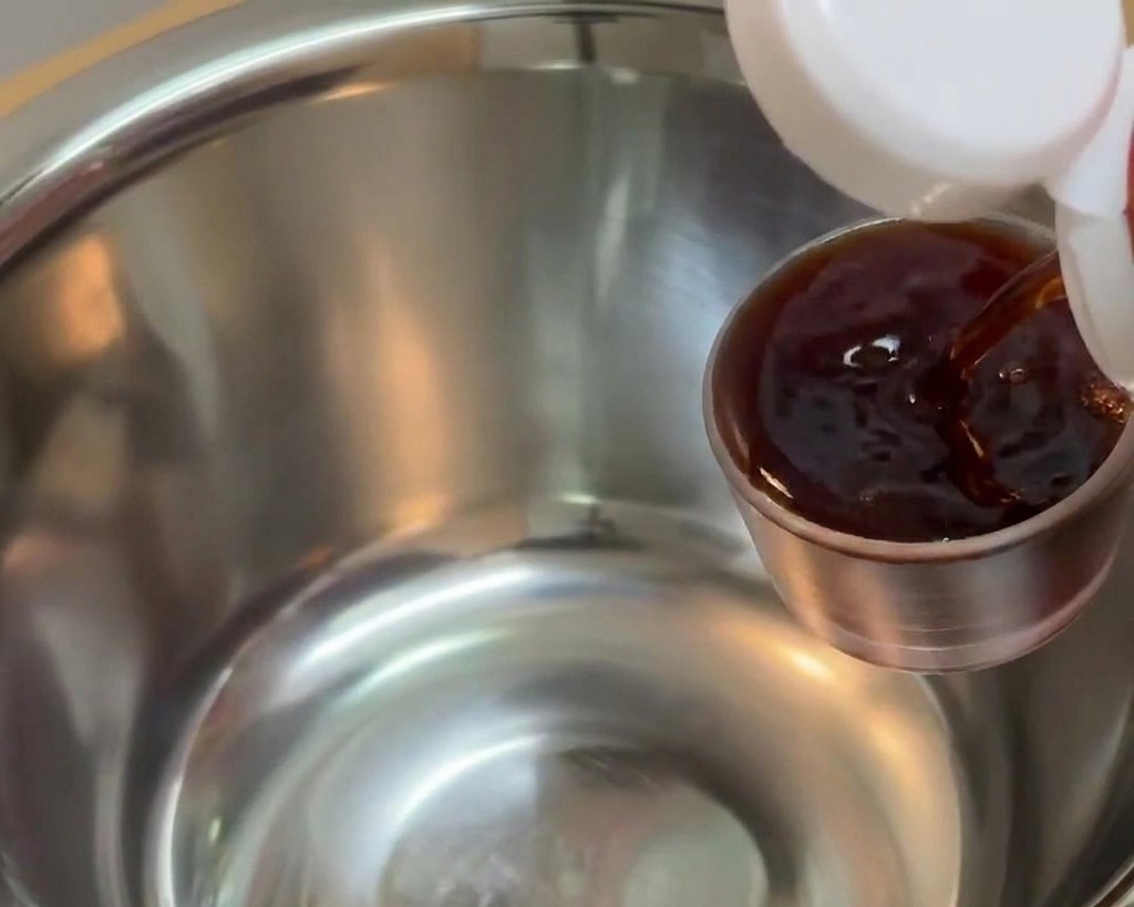 Salsa de soja oscura vertiéndose desde una pequeña taza medidora de metal en un bol para mezclar de acero inoxidable más grande.
