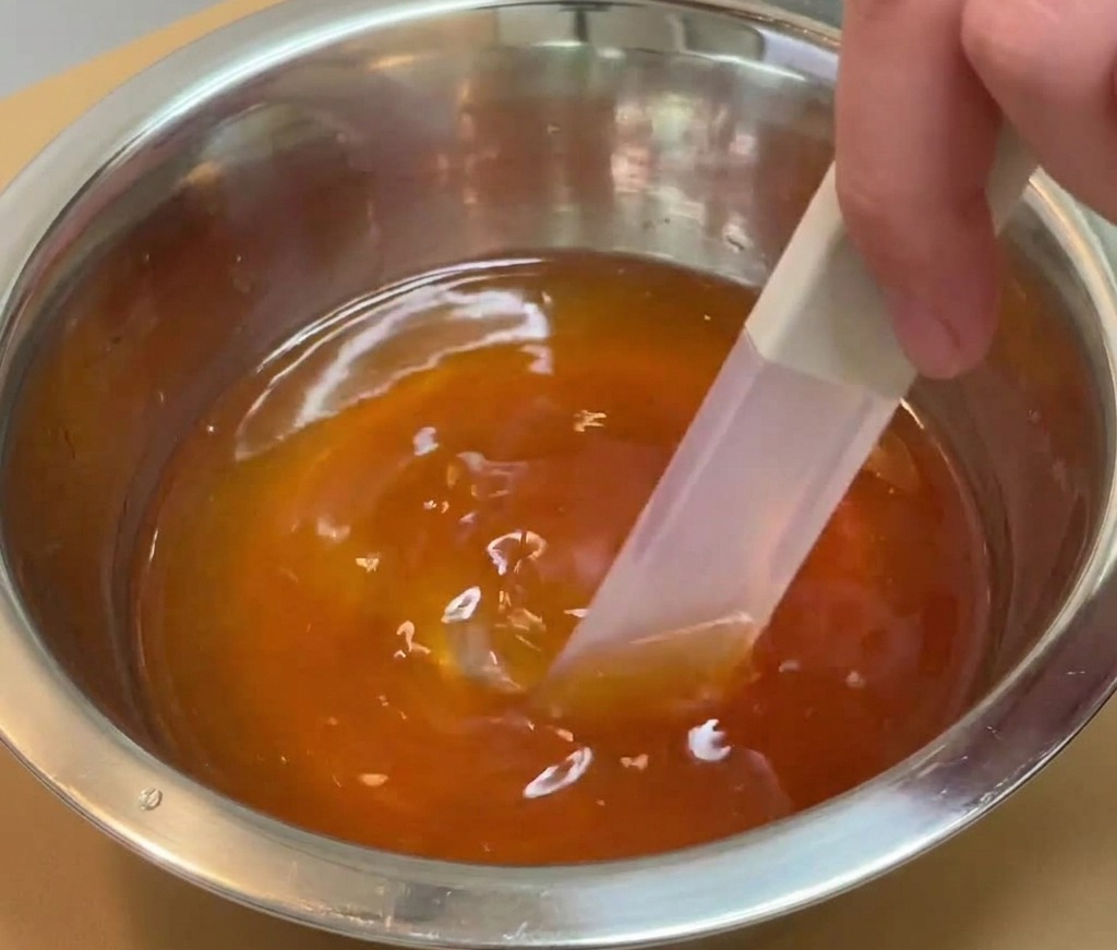 Una mano usando una espátula para remover un líquido de condimento oscuro y translúcido en un bol de acero inoxidable.