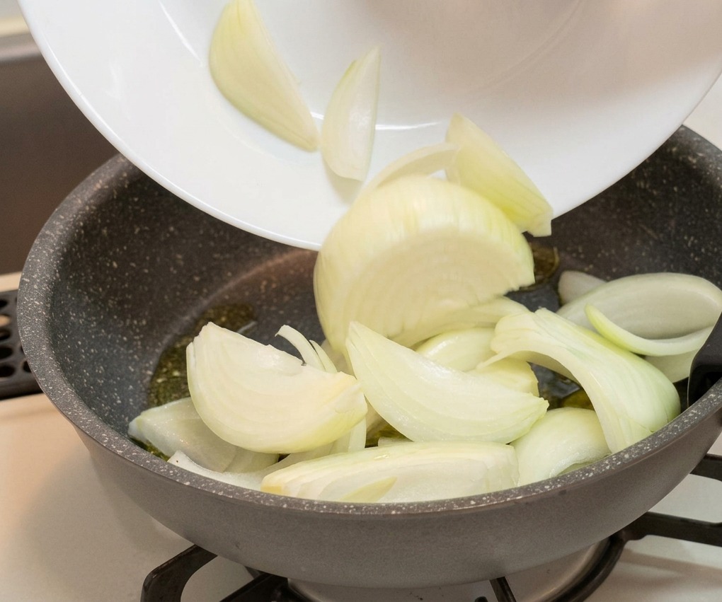 Gajos de cebolla blanca cortados siendo pasados de un plato blanco a una sartén antiadherente gris.