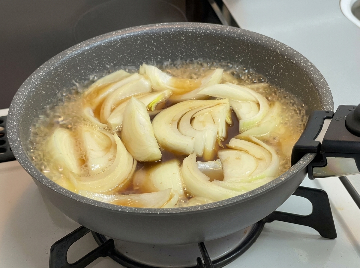 Cebollas cocinándose a fuego lento en un caldo burbujeante de color marrón oscuro dentro de una sartén gris en la estufa.