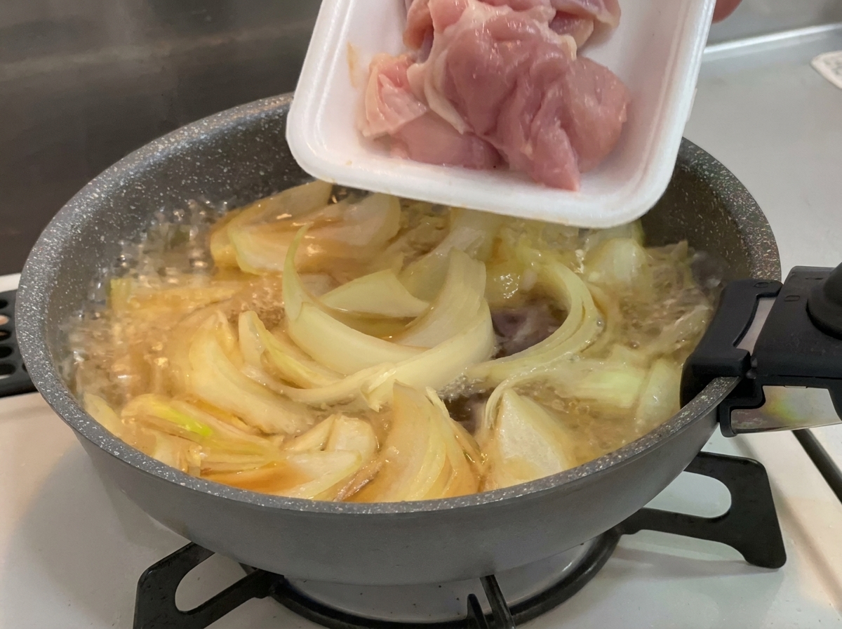 Trozos de contramuslo de pollo crudo de color rosa siendo añadidos desde una bandeja de poliestireno blanco a una sartén con cebollas y caldo hirviendo.