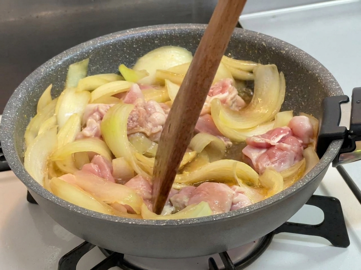 Una espátula de madera removiendo trozos de pollo parcialmente cocidos y cebollas en un líquido oscuro que hierve a fuego lento en una sartén.