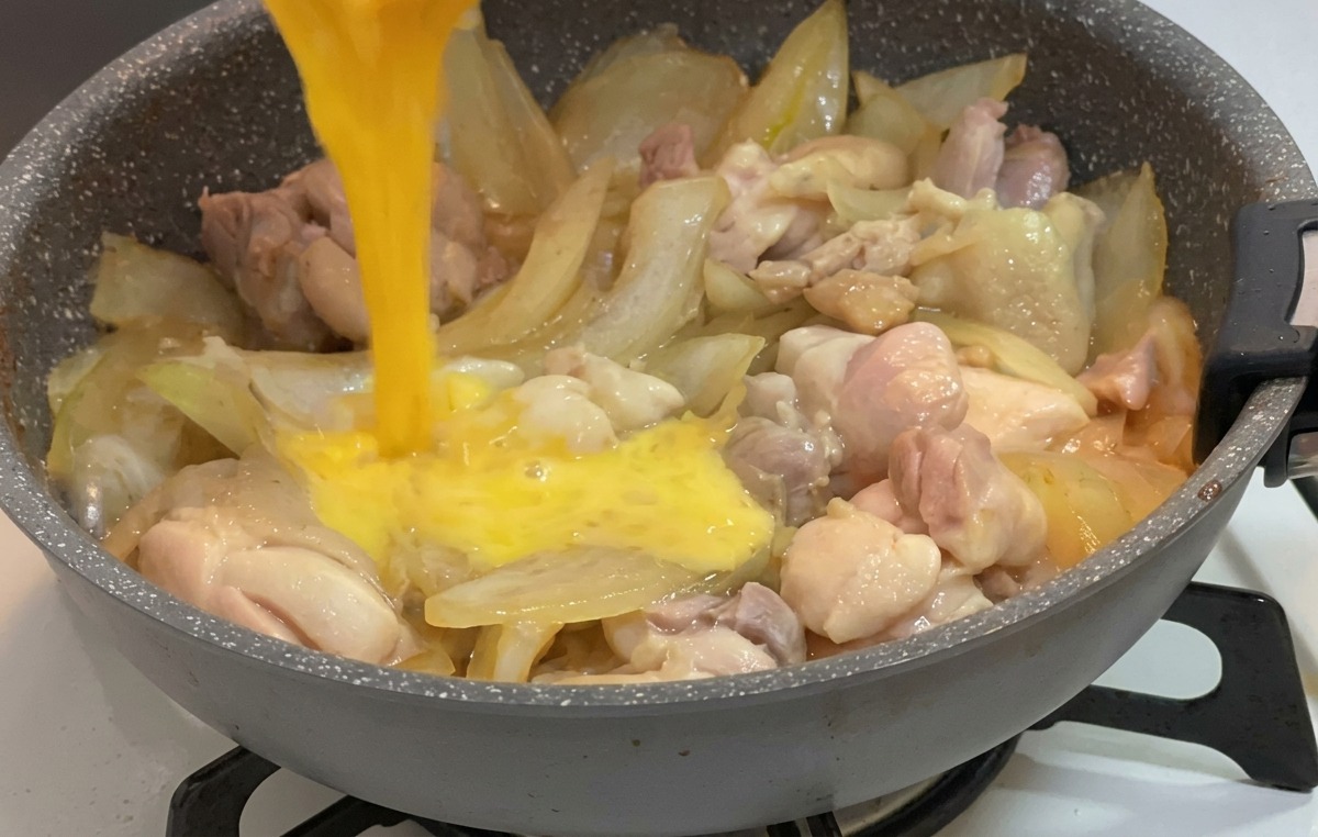 Huevo batido vertiéndose desde un tazón pequeño sobre pollo y cebollas que hierven a fuego lento en una salsa.