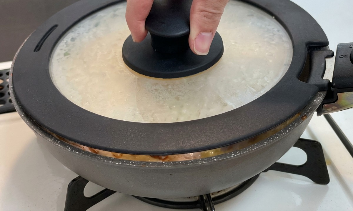 Una mano colocando una tapa de vidrio con un pomo negro sobre una sartén que contiene pollo, cebollas y huevos.