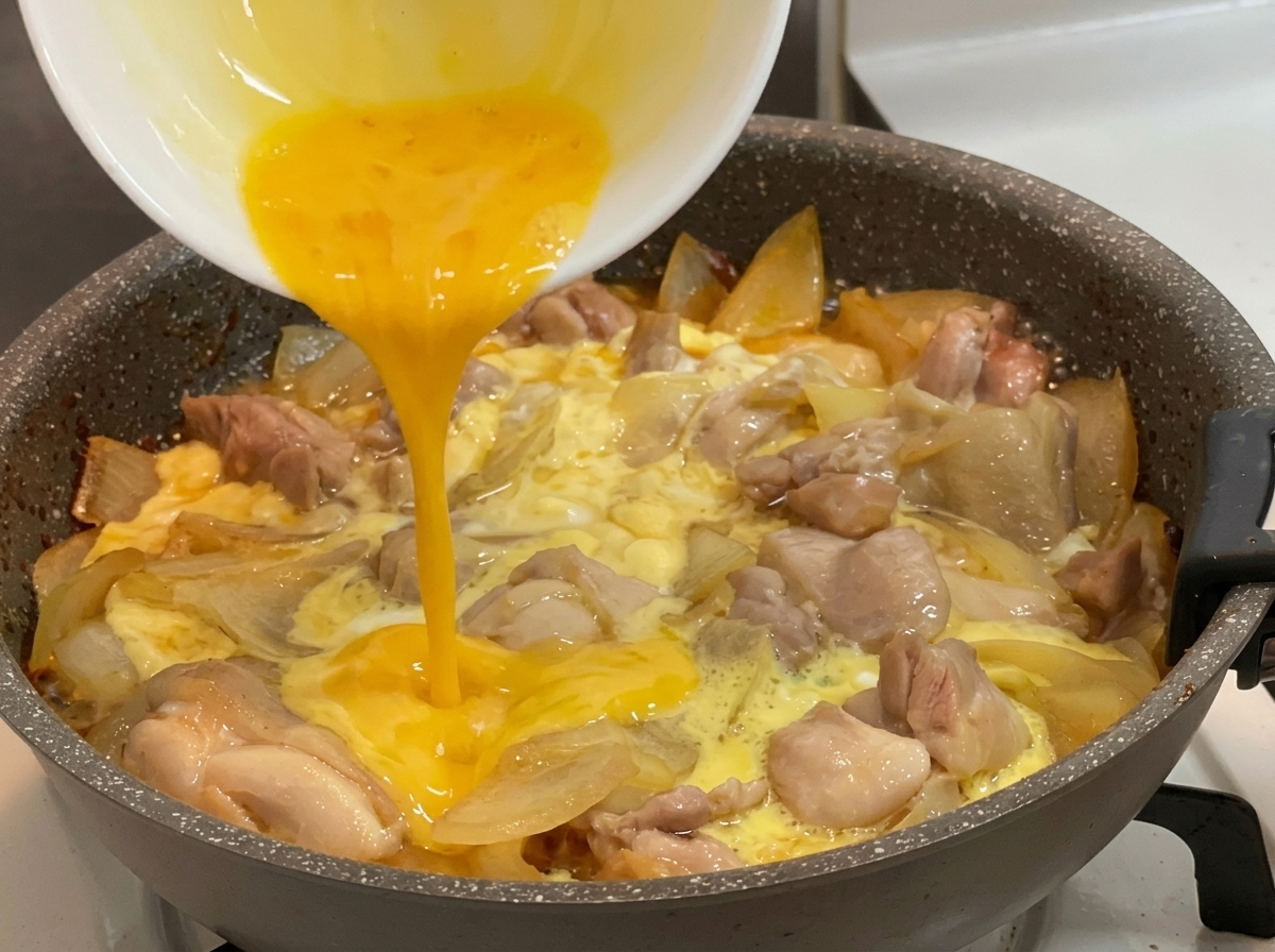 Vertiendo huevos batidos de color amarillo brillante desde un bol blanco en una sartén caliente llena de trozos de pollo hirviendo, cebollas cortadas y huevos parcialmente cuajados.