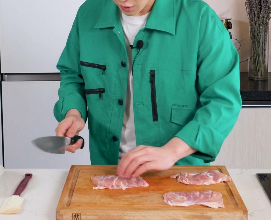 Un cuchillo haciendo cortes superficiales en la carne de contramuslos de pollo en una tabla de picar.