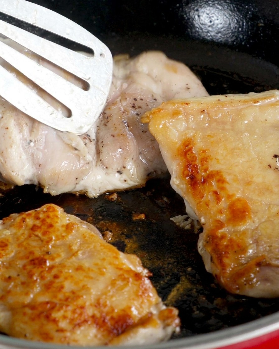 Contramuslos de pollo volteados en una sartén, con la piel dorada hacia arriba, mientras se cocina el lado de la carne.