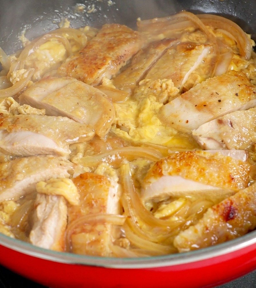 Trozos de pollo dorado entre cebollas translúcidas en una sartén roja, con huevo crudo fresco vertiéndose encima para crear una salsa cremosa.