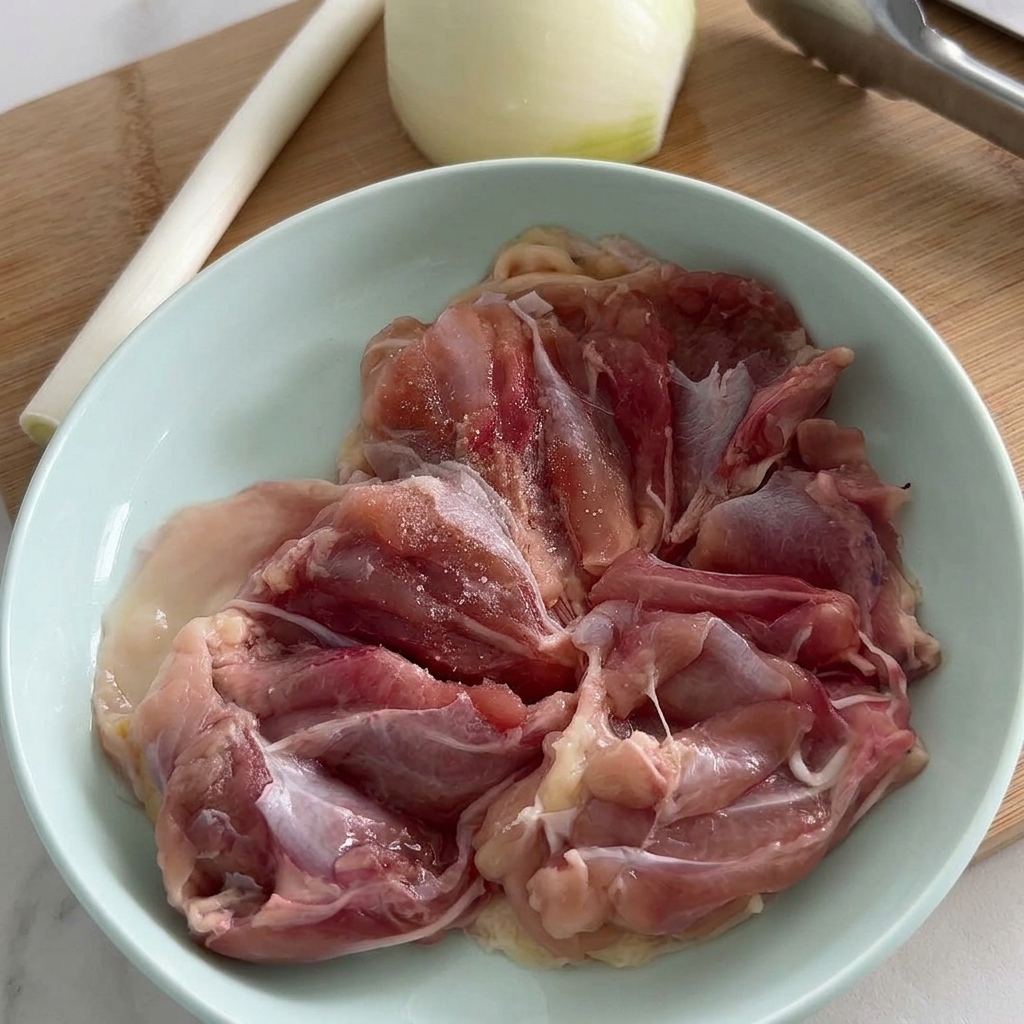 Filetes de muslo de pollo crudos en un tazón azul claro siendo sazonados con sal y pimienta negra recién molida.