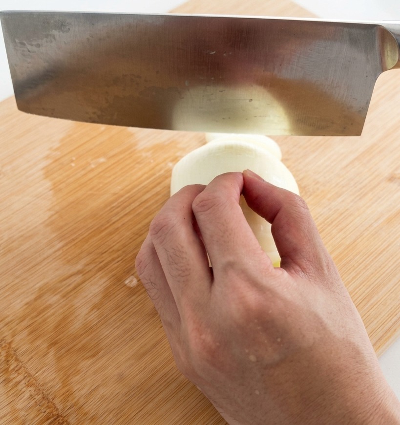 Una mano usando un gran cuchillo de acero inoxidable para cortar una cebolla blanca en tiras finas sobre una tabla de cortar de bambú.