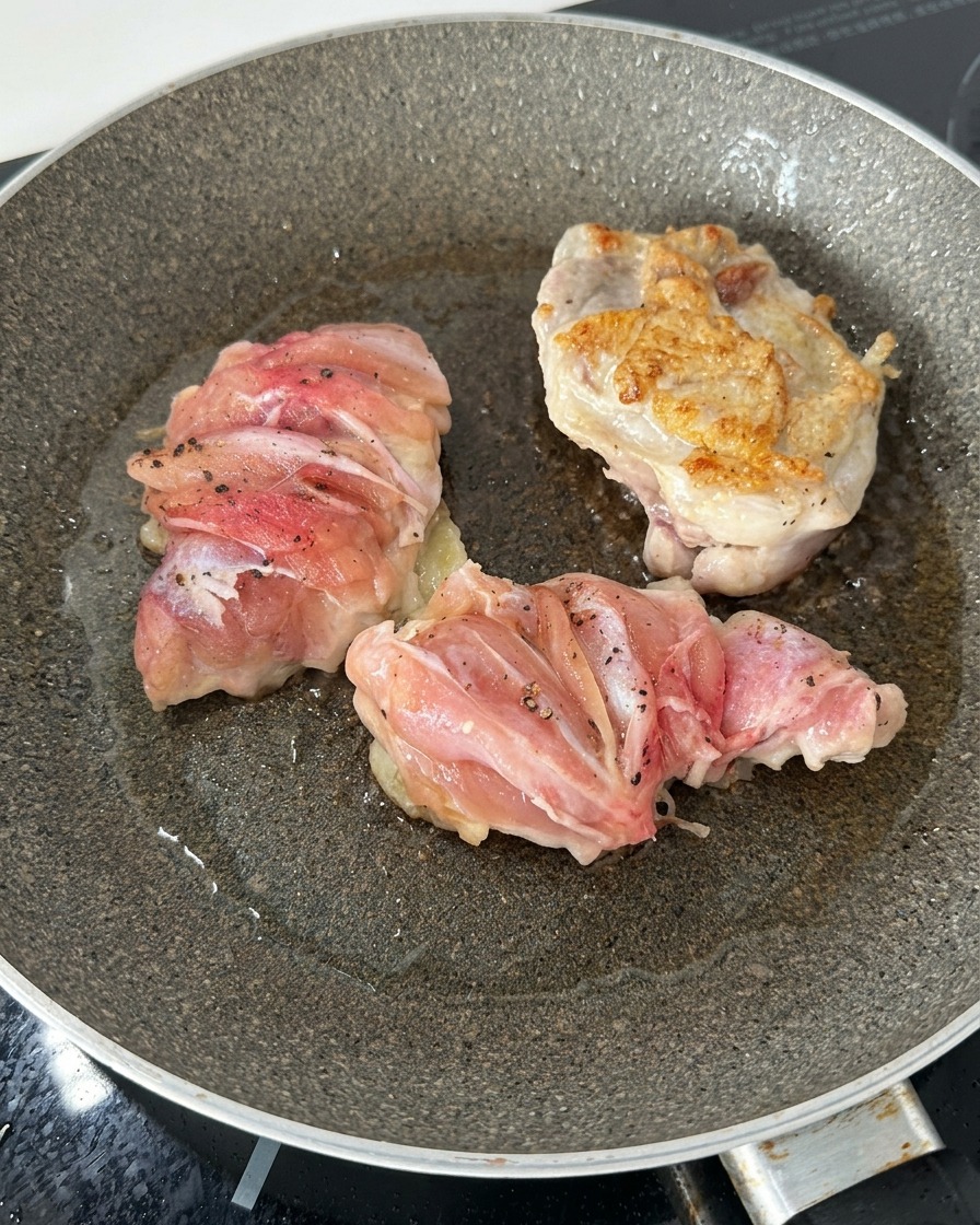 Tres muslos de pollo en una sartén, uno de ellos volteado mostrando una superficie dorada y crujiente por el sellado.