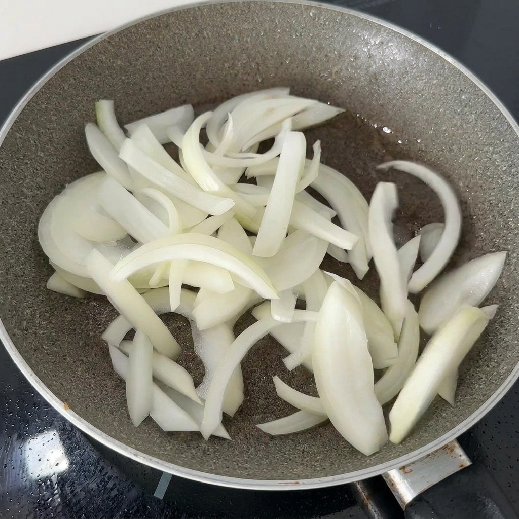 Tiras finas de cebolla blanca salteándose en una sartén oscura con el aceite residual hasta que se ablanden.