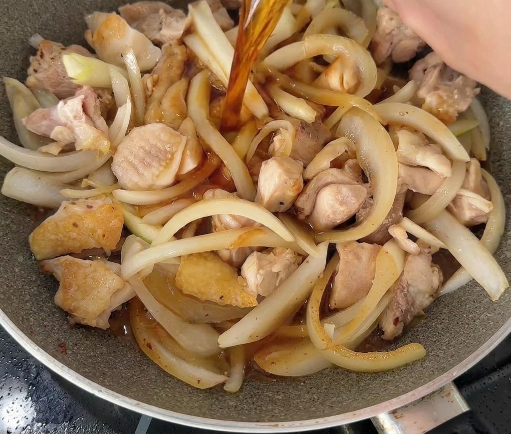Un líquido oscuro y sabroso vertiéndose desde un tazón blanco sobre una sartén llena de trozos de pollo y cebolla.