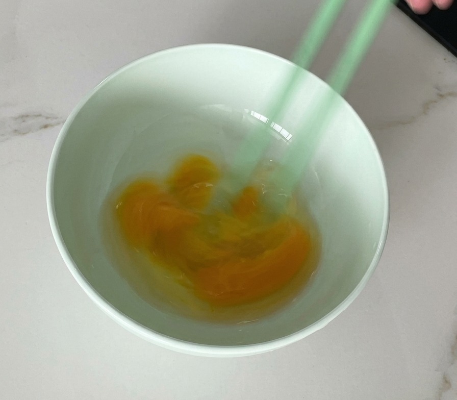 Un par de palillos verdes batiendo huevos crudos en un tazón verde claro, mostrando una mezcla veteada de amarillo y blanco.