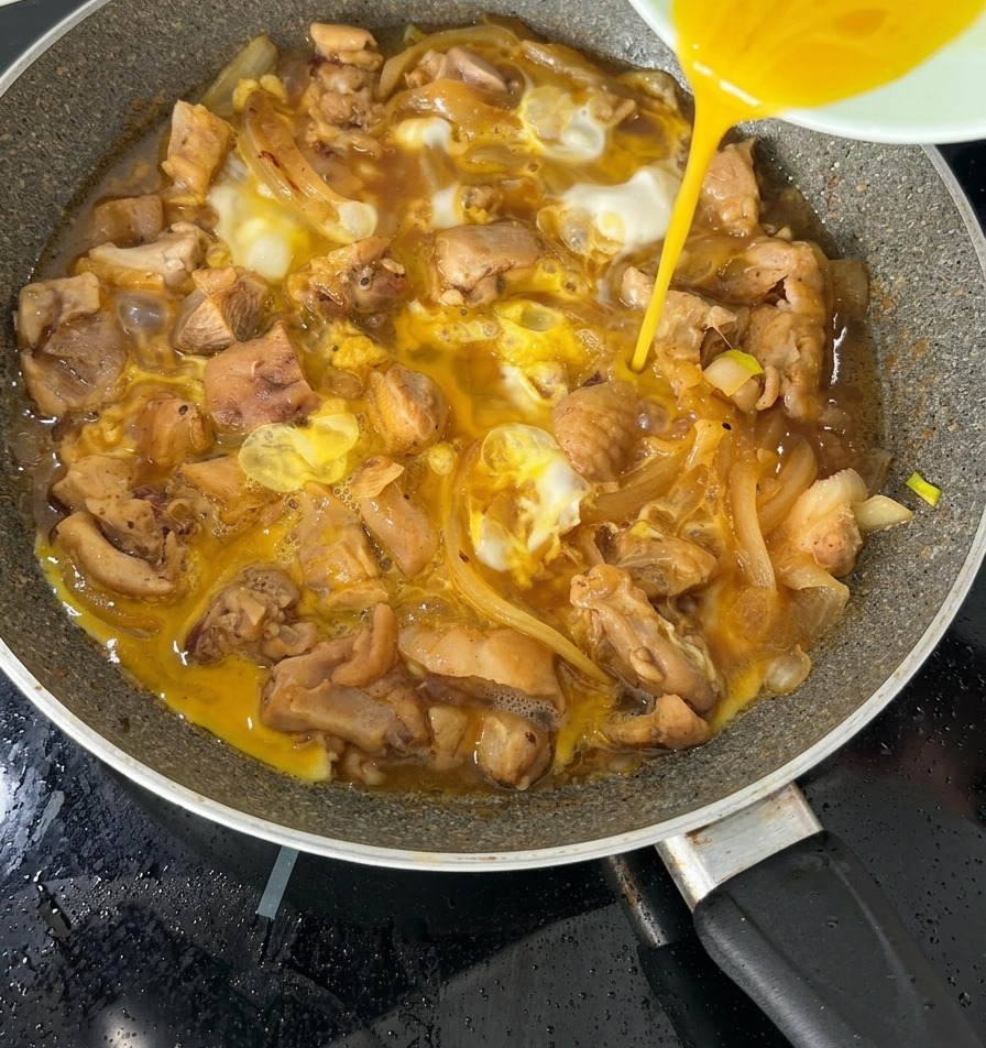 La segunda mitad de la mezcla de huevo batido vertiéndose sobre un Oyakodon casi terminado en una sartén.