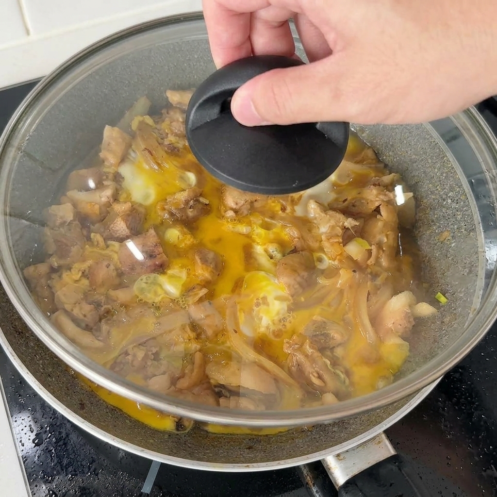 Una mano colocando una tapa de vidrio sobre una sartén de pollo y huevos para cocinar al vapor la última adición de la mezcla de huevo.