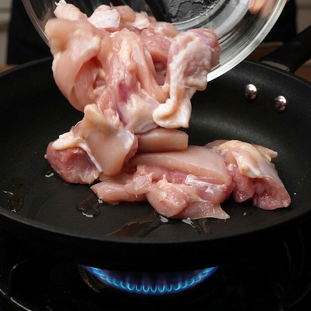 Trozos de pollo crudo cayendo desde un bol de vidrio a una sartén caliente con una llama de gas azul visible debajo.