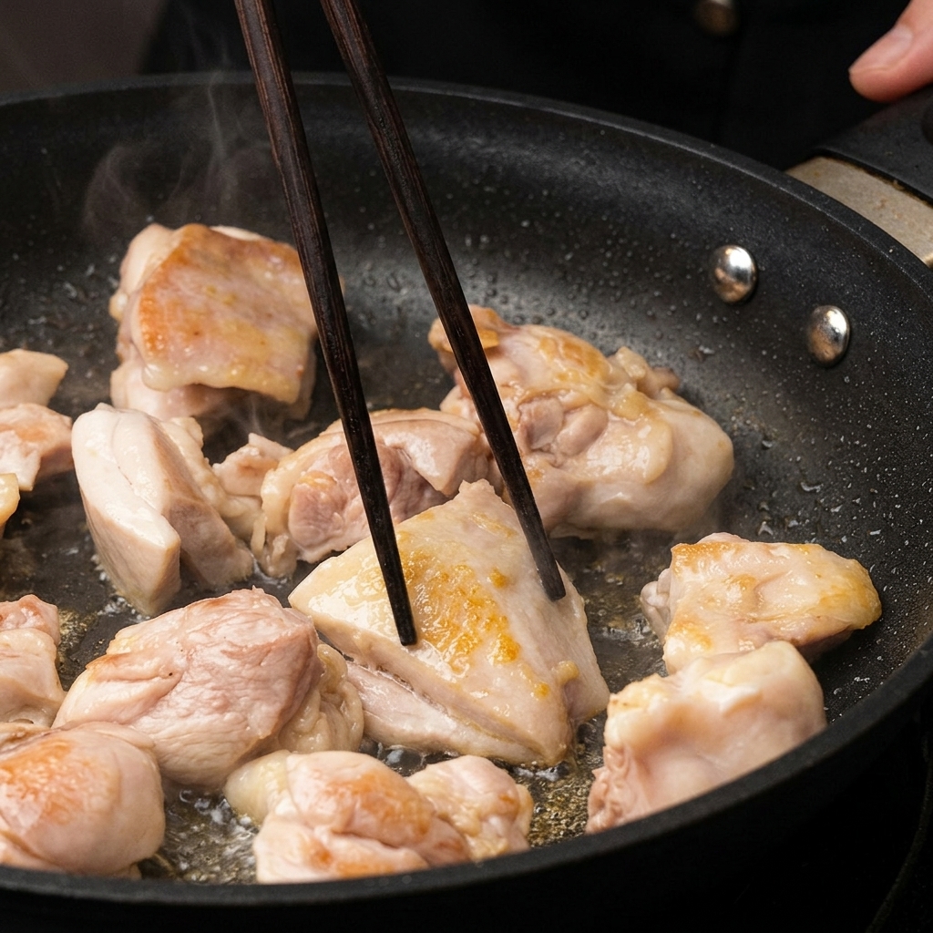 Trozos de pollo dorándose en una sartén, siendo volteados con palillos de madera oscura.